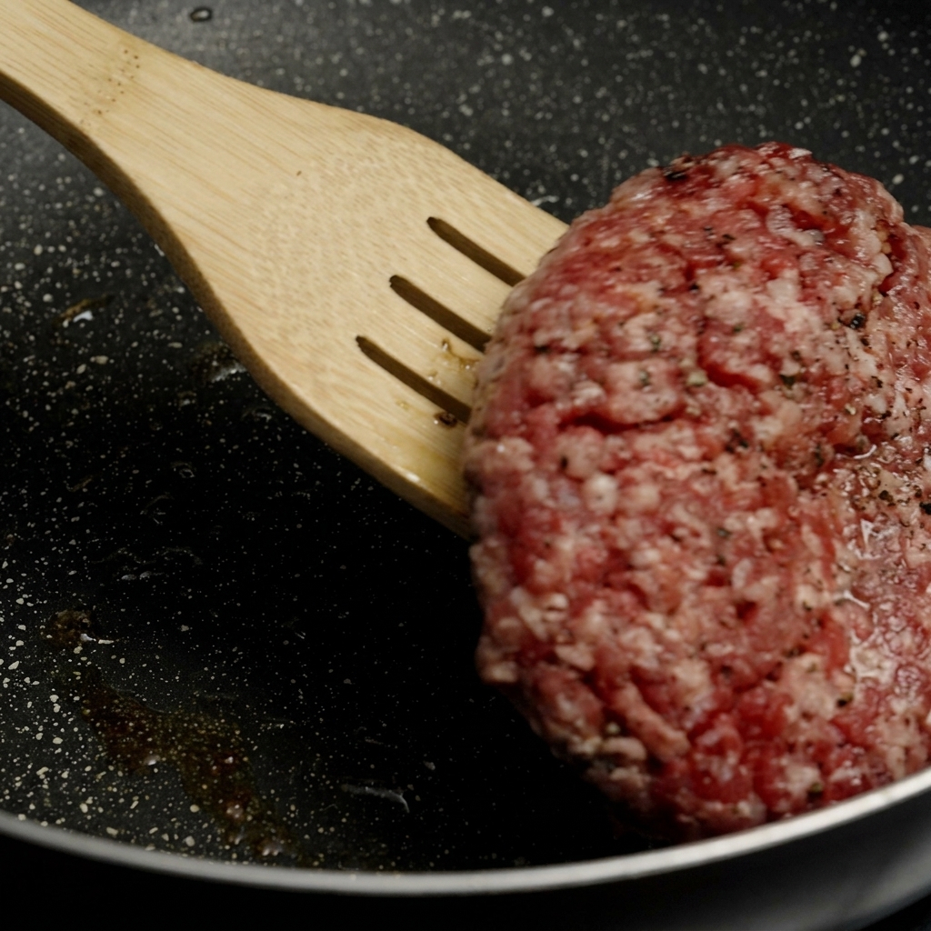 熱いフライパンの中で、こんがりと焼き色のついた分厚いパティを木製のフライ返しで裏返している様子。