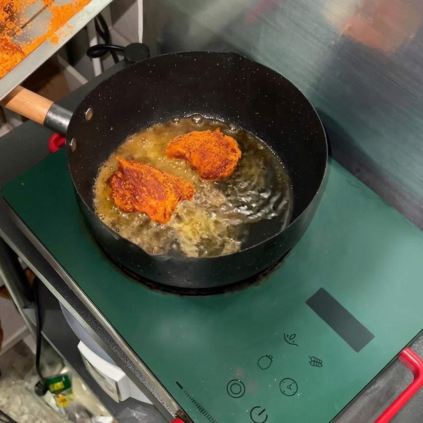 IHコンロの上に置かれた黒いフライパンの中で、スパイスをまぶした豚肉を多めの油で焼いている様子。