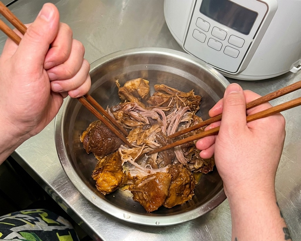 金属製のボウルの中で、柔らかく煮込まれた豚肉の塊を菜箸を使って細かくほぐしている様子。