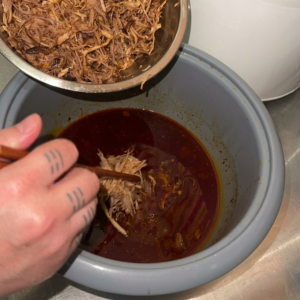 細かくほぐした豚肉を、濃厚な赤い煮汁が残った炊飯器の釜に戻し入れている様子。