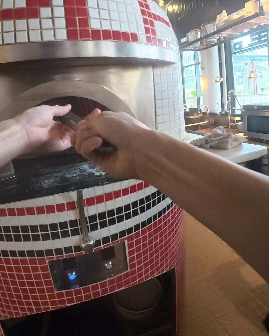 金属製ピールを使って、焼き上がった熱々のピザをオーブンから取り出している様子。