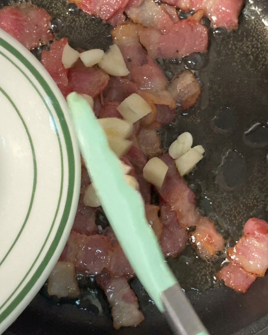 カリカリに炒められたベーコンが入ったフライパンに、みじん切りにしたにんにくを加えている様子。
