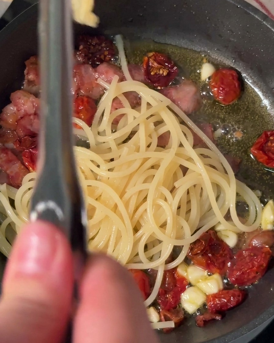 ゆで上がったスパゲッティをトングで挟み、ベーコン、にんにく、ドライトマトが入った熱いフライパンへ直接移している様子。