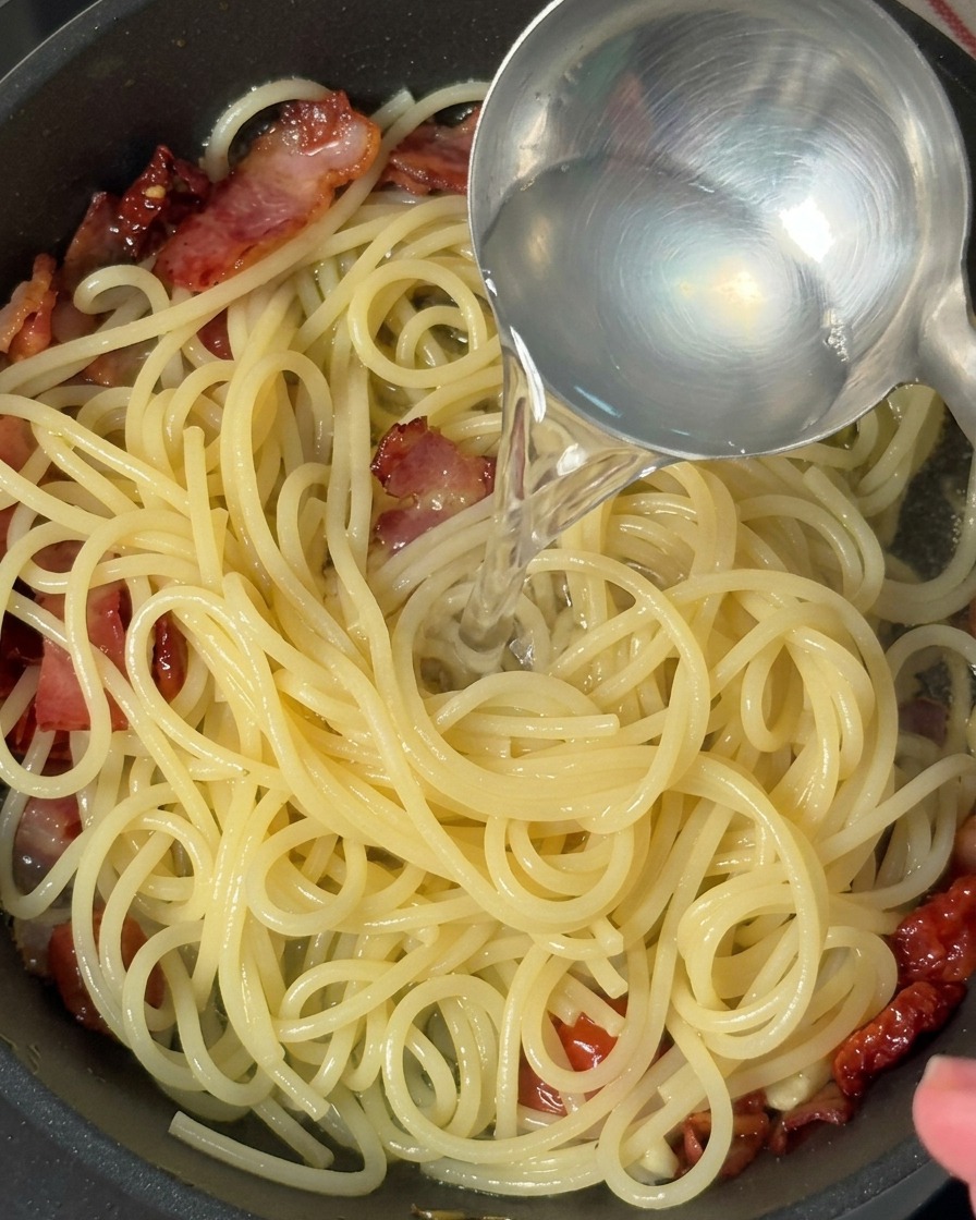 スパゲッティ、ベーコン、トマトが入ったフライパンに、金属製のお玉で透明なゆで汁を注いでいる様子。