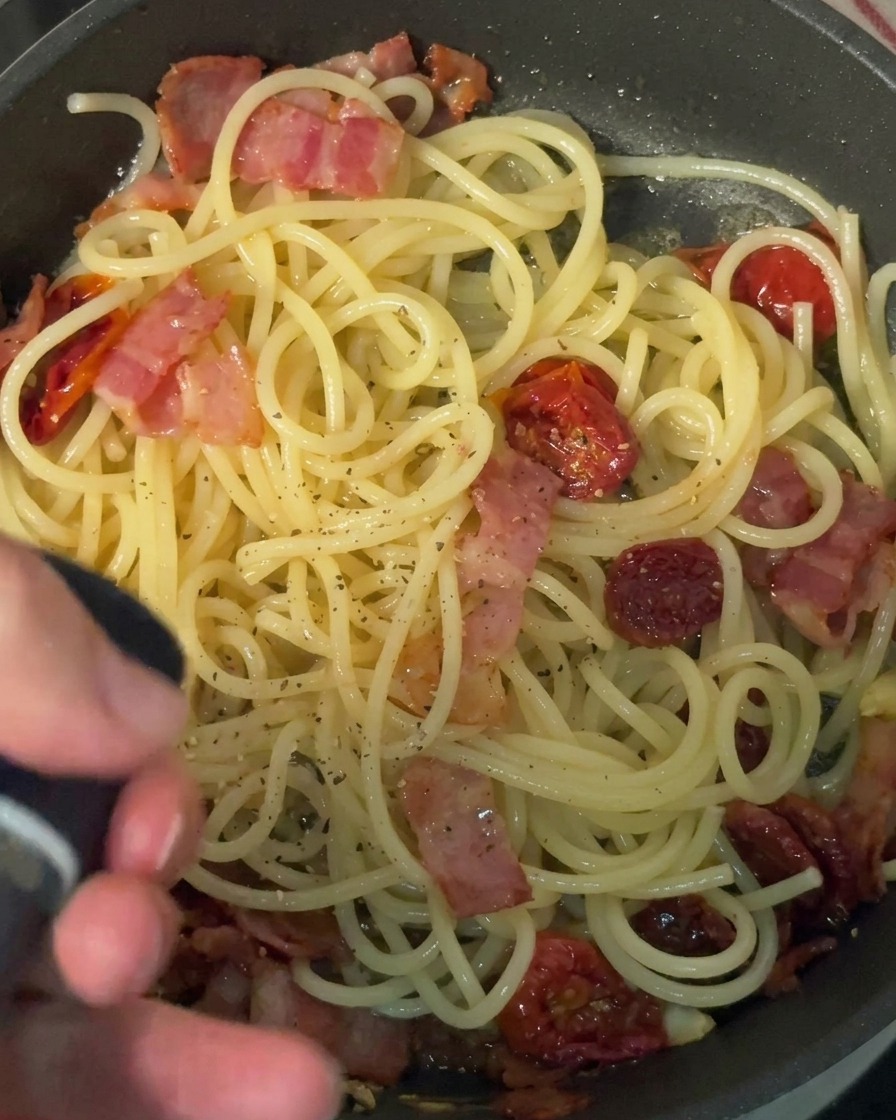 ペッパーミルを使って、パスタと具材が入ったフライパンに粗挽きの黒こしょうを挽いている手元の様子。