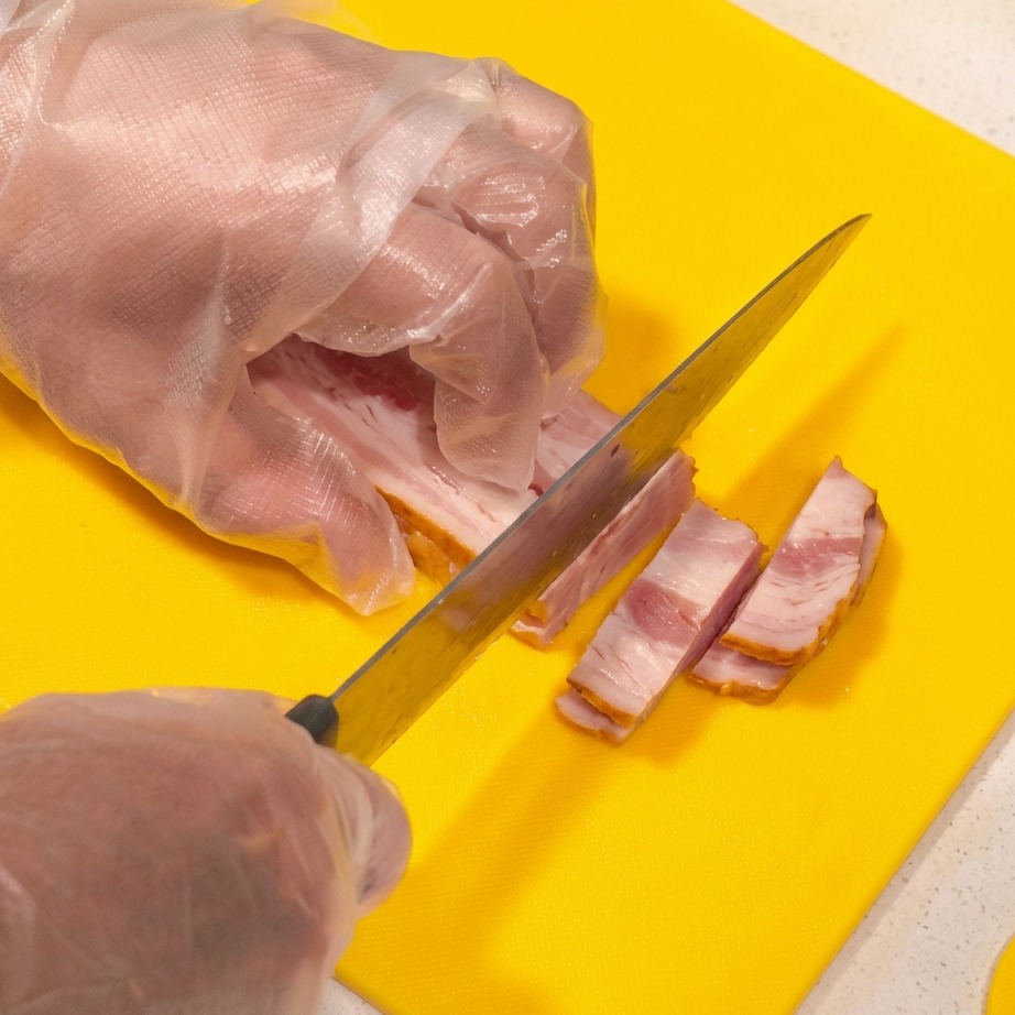 明るい黄色のまな板の上で、生の厚切りベーコンを棒状に切っている様子。