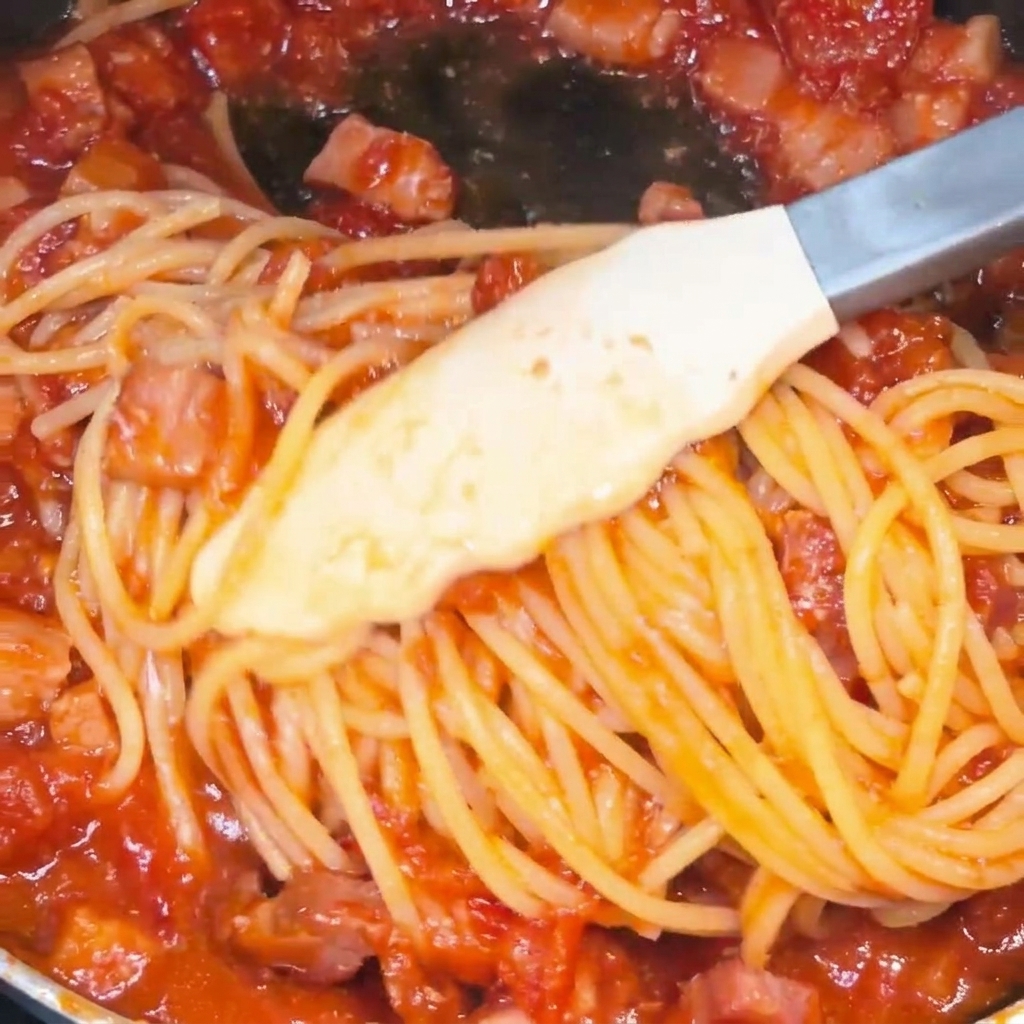 フライパンの中でトングを使ってスパゲッティを和え、鮮やかな赤いトマトとベーコンのソースをしっかり絡ませている様子。