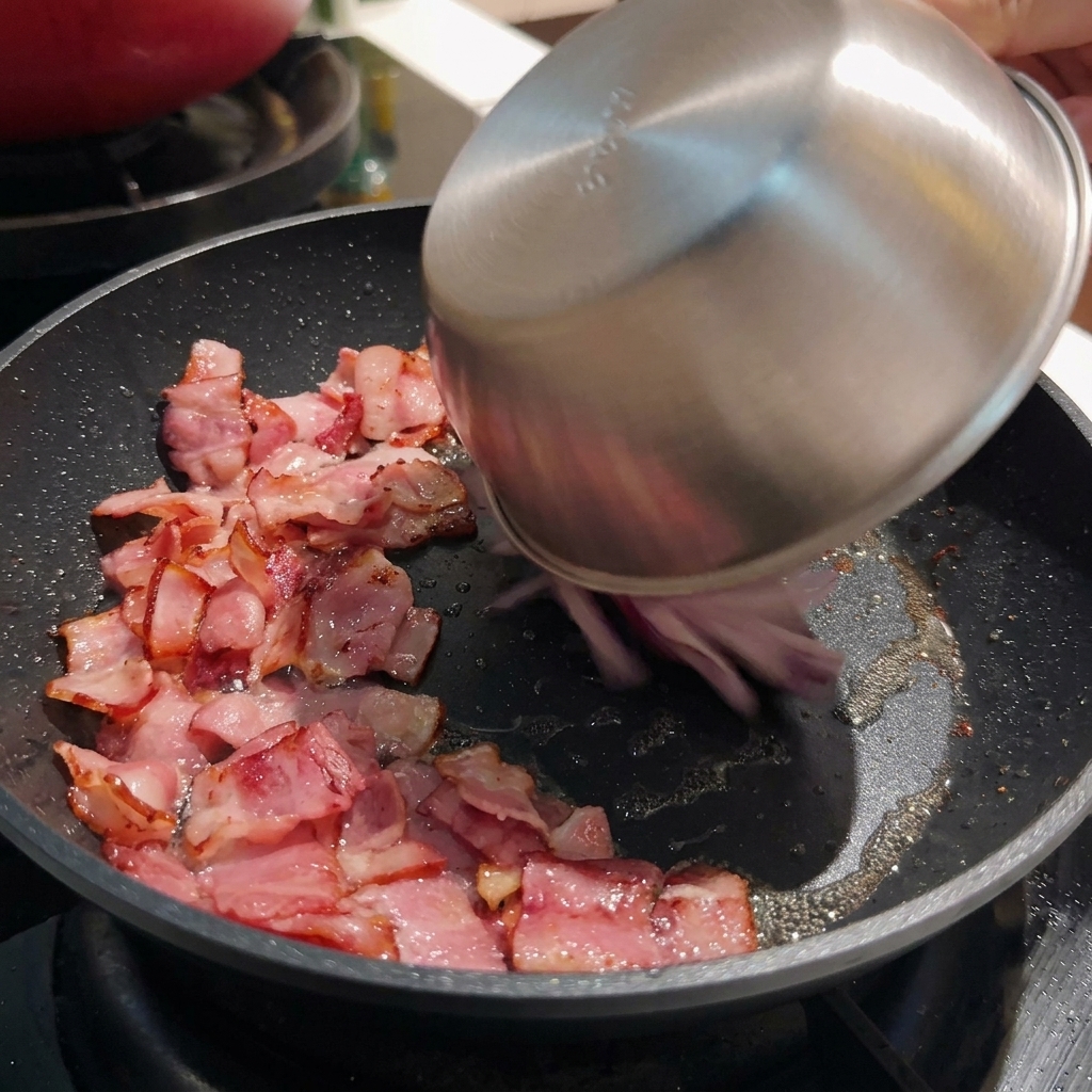 ボウルからスライスした赤玉ねぎを、カリカリのベーコンが入ったフライパンに入れている様子。