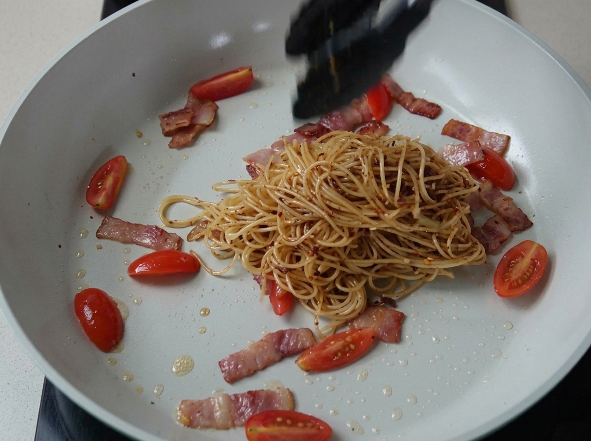 茹でたパスタをベーコンとトマトの入ったフライパンに加え、合わせている様子。