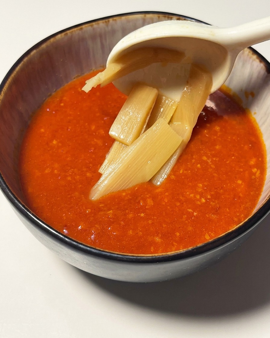鮮やかなトマト海老のつけ汁が入った器に、スプーンでメンマを盛り付けている様子。