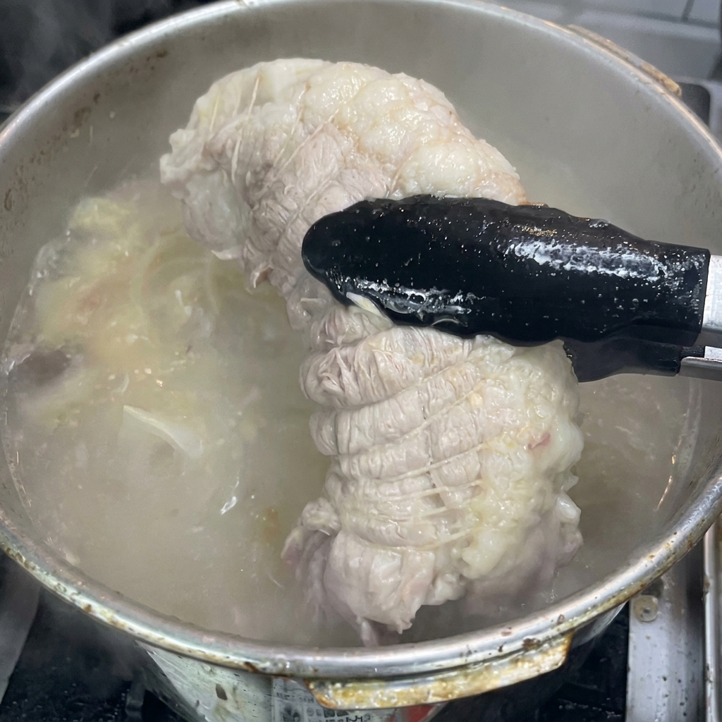 トングを使って煮上がった豚肩ロースを取り出し、タレの容器へ入れる様子。