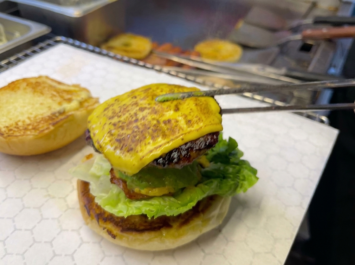 トングを使って、チーズを乗せて炙ったビーフパティとパイナップルリングをバーガーのベースに乗せている様子。