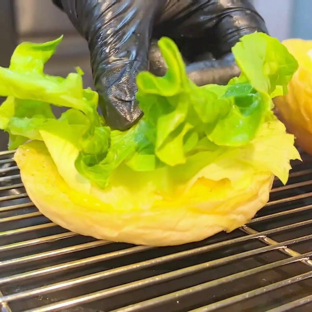 下のバンズの上に、レタス、トマト、チーズバーガー、ベーコンが順に積み重なっている組み立て中のバーガー。