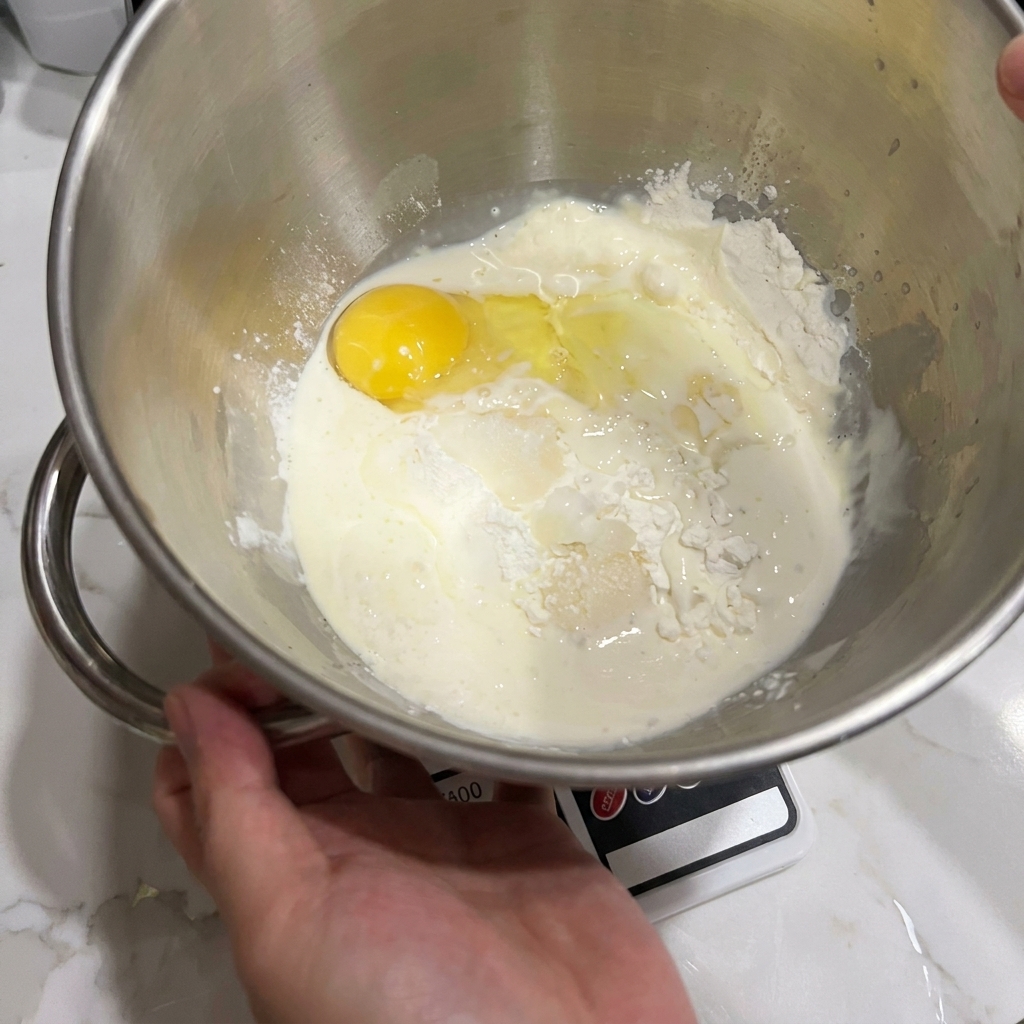 スタンドミキサーのボウルに小麦粉、卵、牛乳が入っており、混ぜる直前の状態。