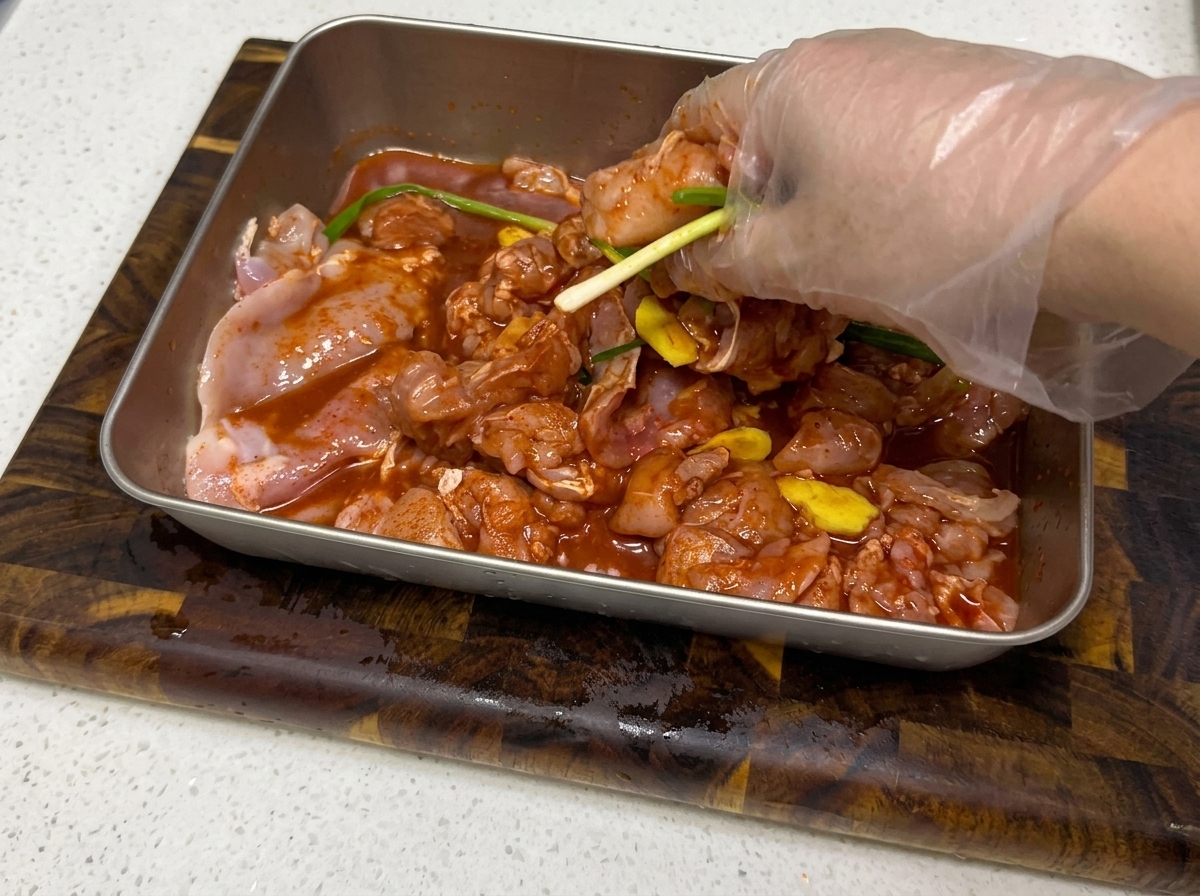 手袋をした手で、金属トレイに入った鶏肉とマリネ液を混ぜ合わせている様子。