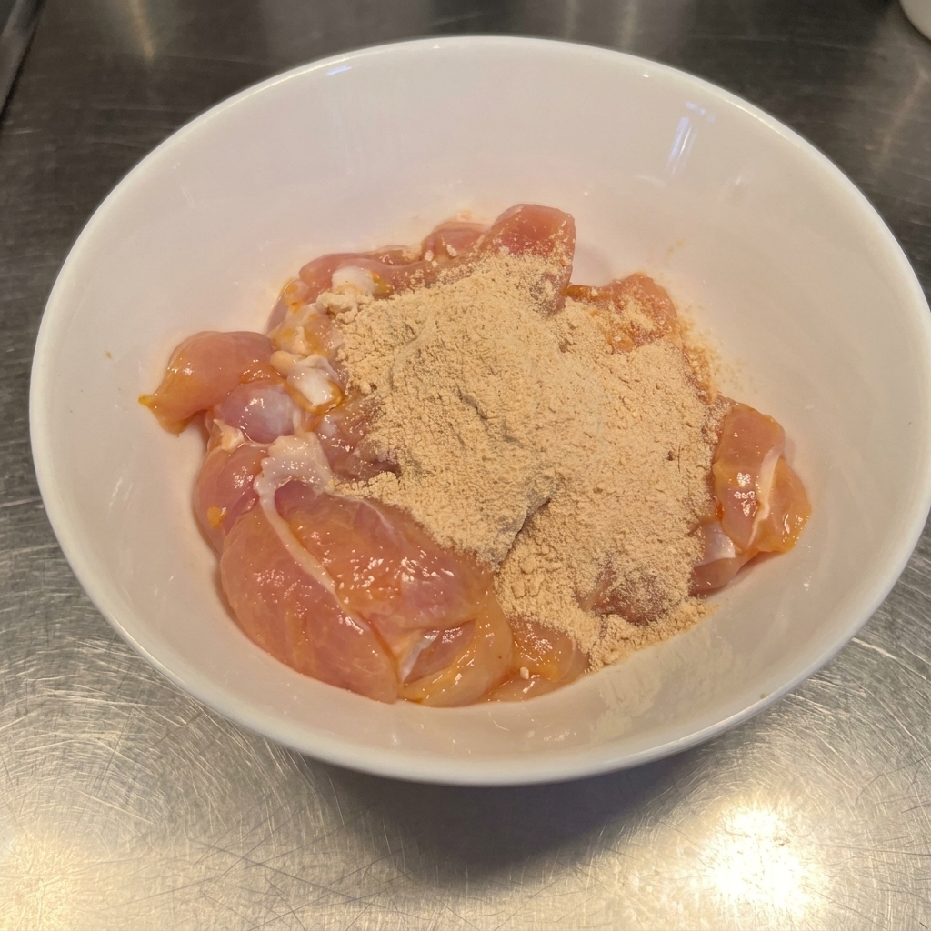ボウルに入った生の鶏もも肉に、赤茶色のチリパウダーとガーリックの調味料がまぶされている様子。