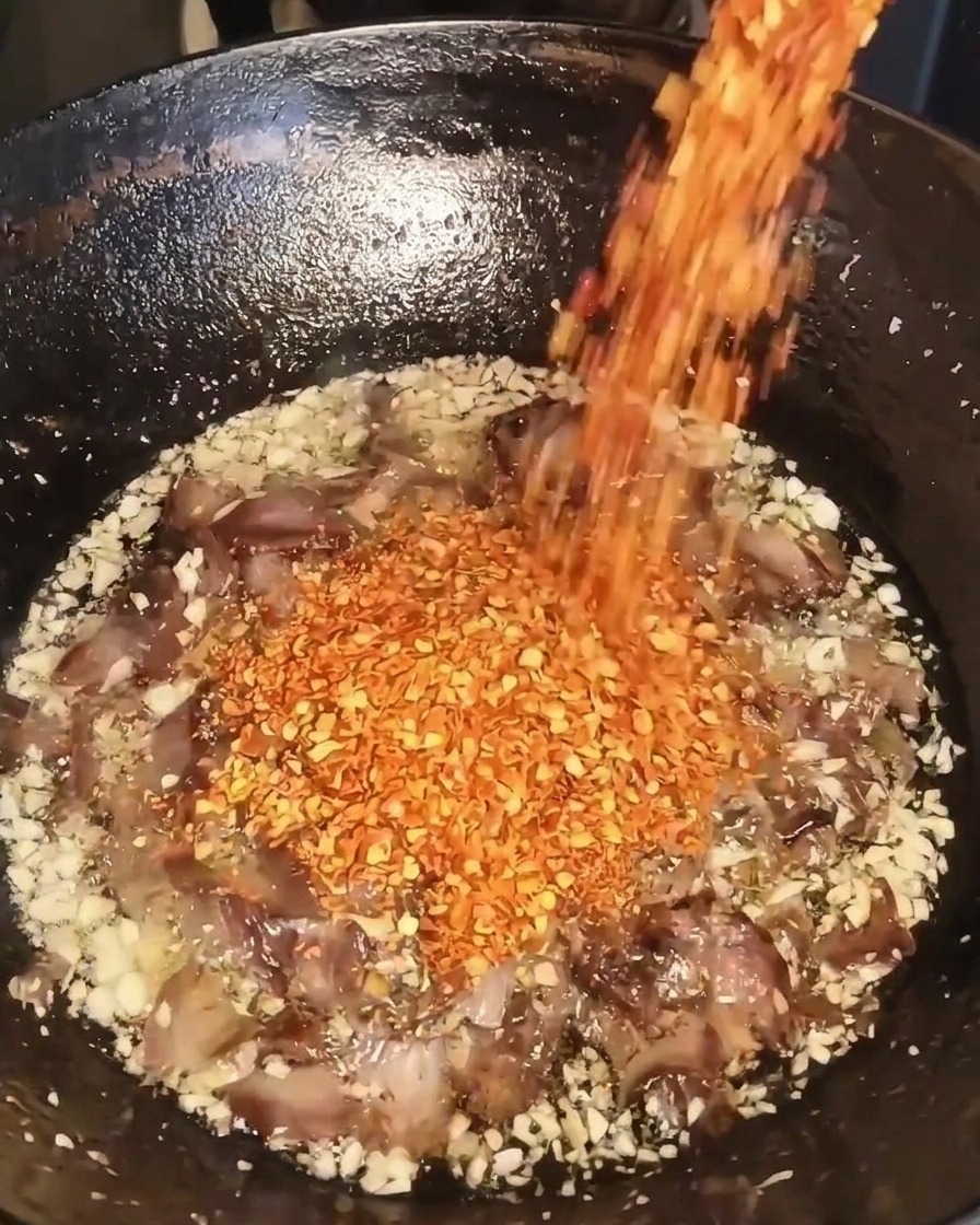 ニンニク、牛肉、油が入った中華鍋に、容器から粗挽き赤唐辛子を注ぎ入れている様子。