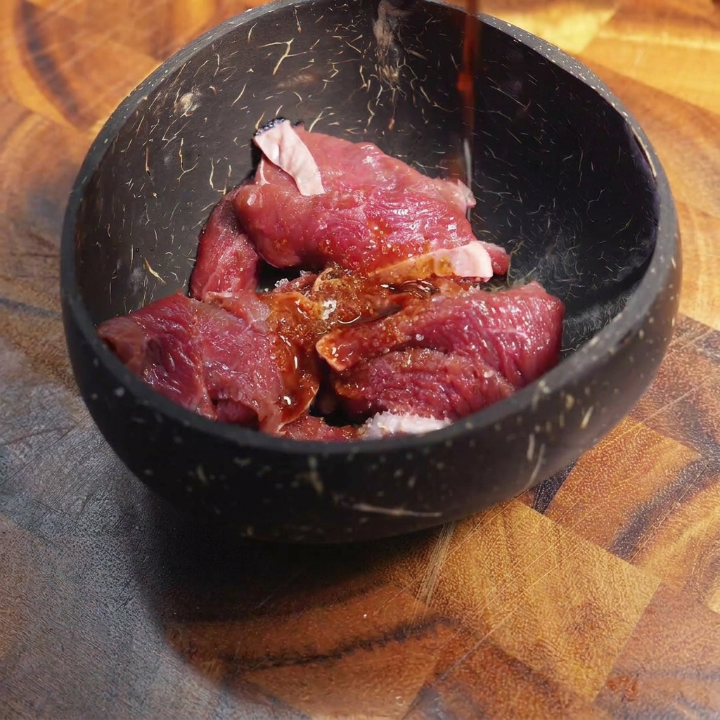 黒いボウルに入った薄切りの生牛肉に、濃い色の調味液をかけている様子。