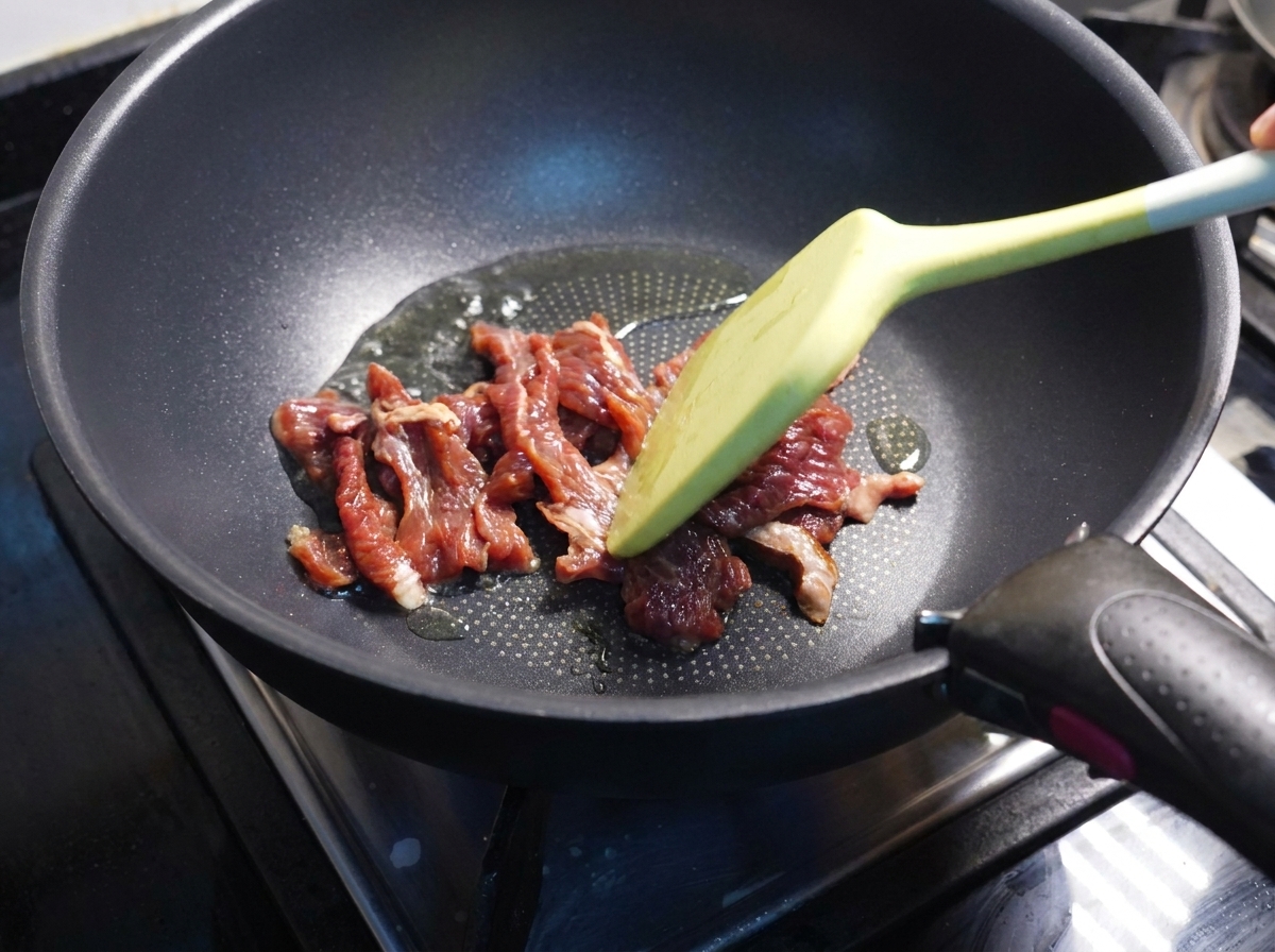 黒いフライパンで、半分ほど火の通った牛肉をヘラで炒めている様子。