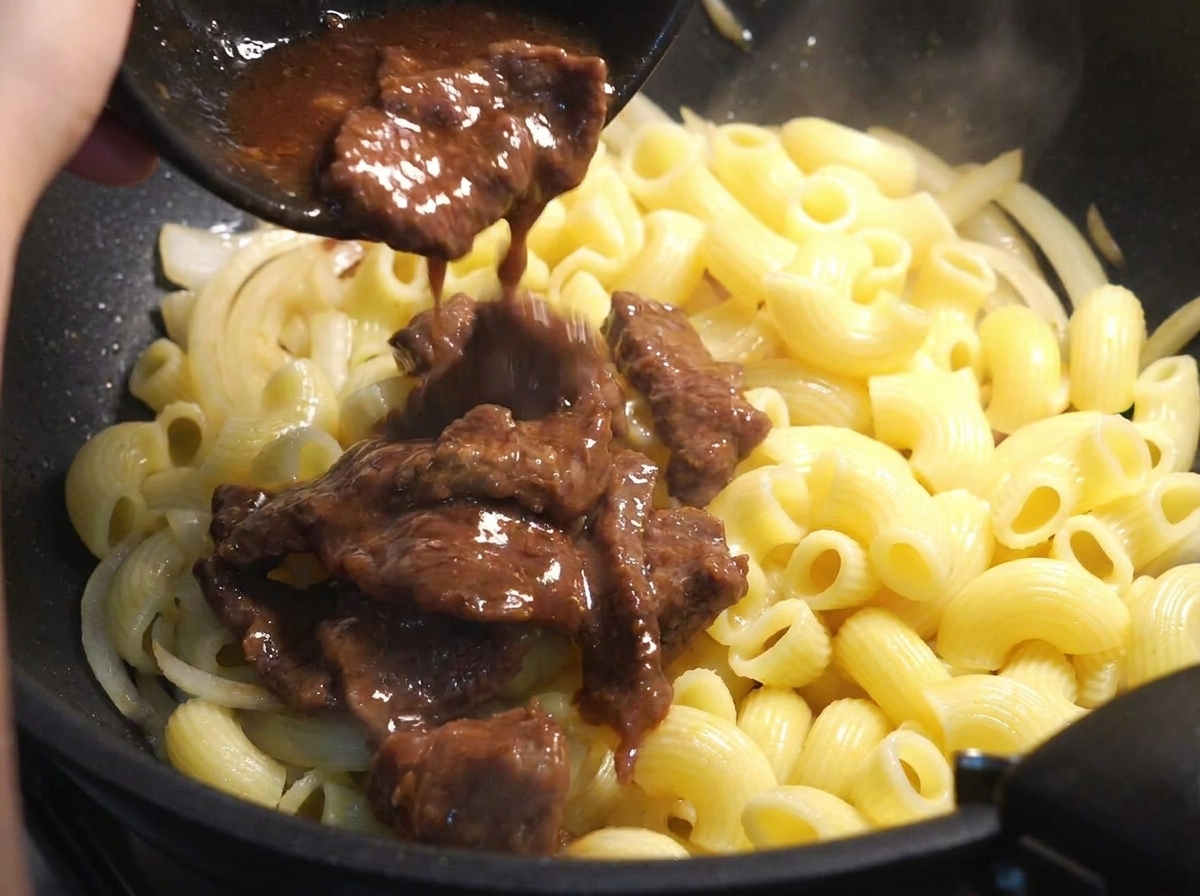 マカロニと玉ねぎの入ったフライパンに、半分火の通った牛肉と漬け汁を戻し入れている様子。