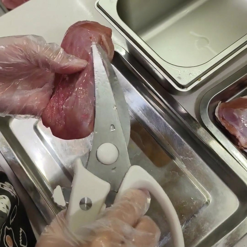 手袋をした手で生の鴨肉を持ち、キッチンバサミで白い脂肪の小さな塊を切り取っている様子。