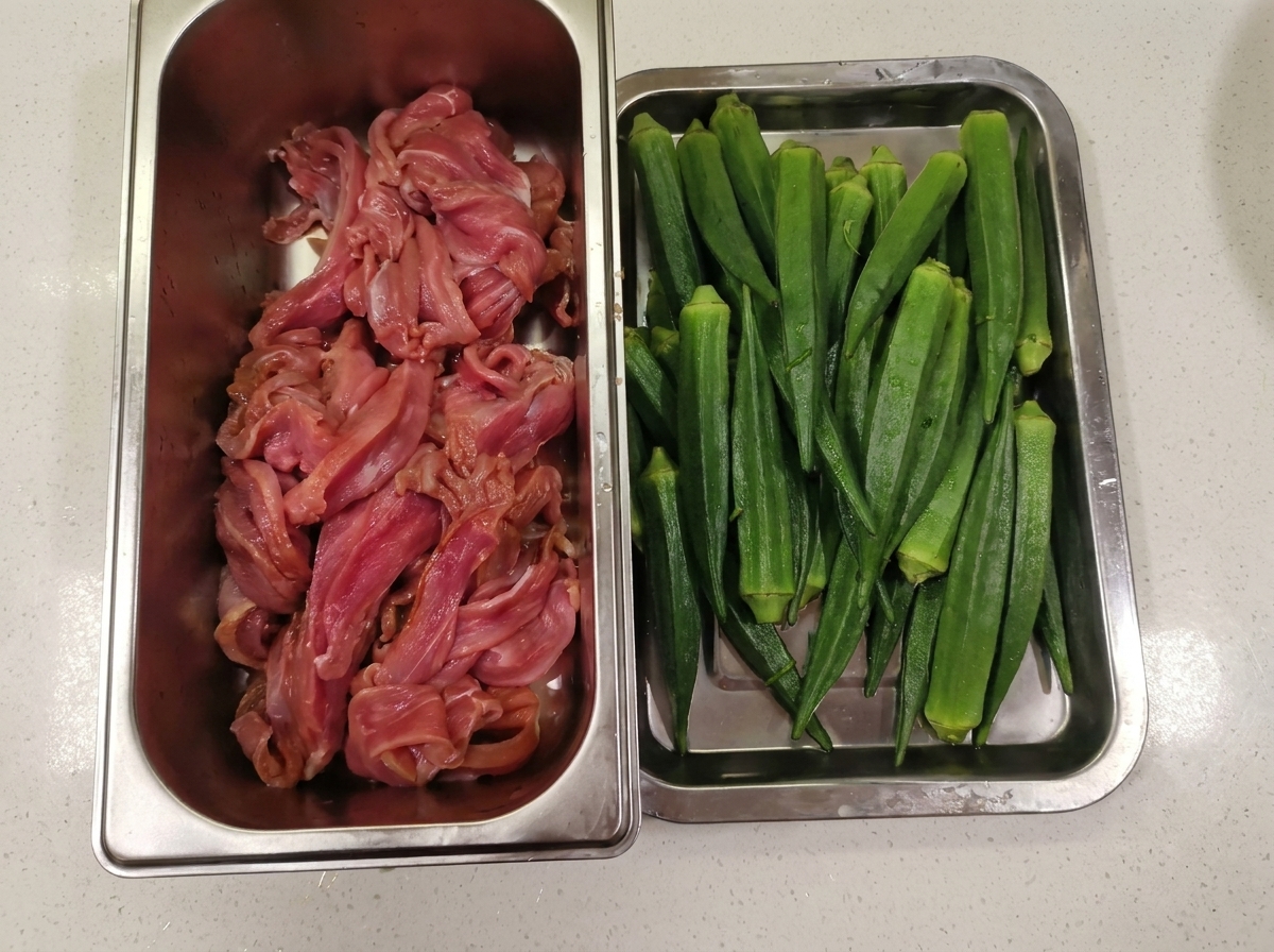 薄切りにした生の鴨肉が入った金属製の容器が、綺麗に洗った緑のオクラが並ぶトレイの横に置かれている様子。