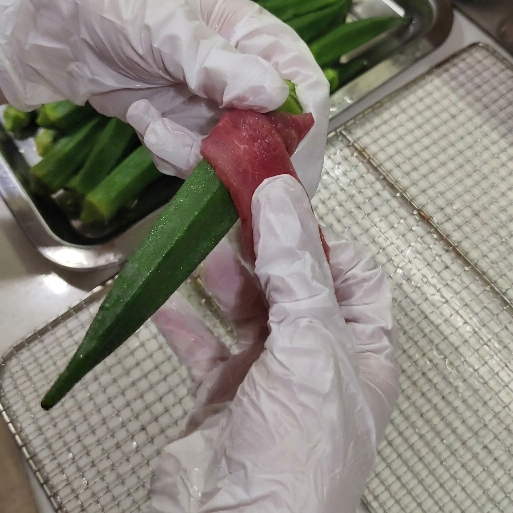手袋をした手で、新鮮な緑のオクラの中心に薄切りにした生の鴨肉をしっかりと巻きつけている様子。