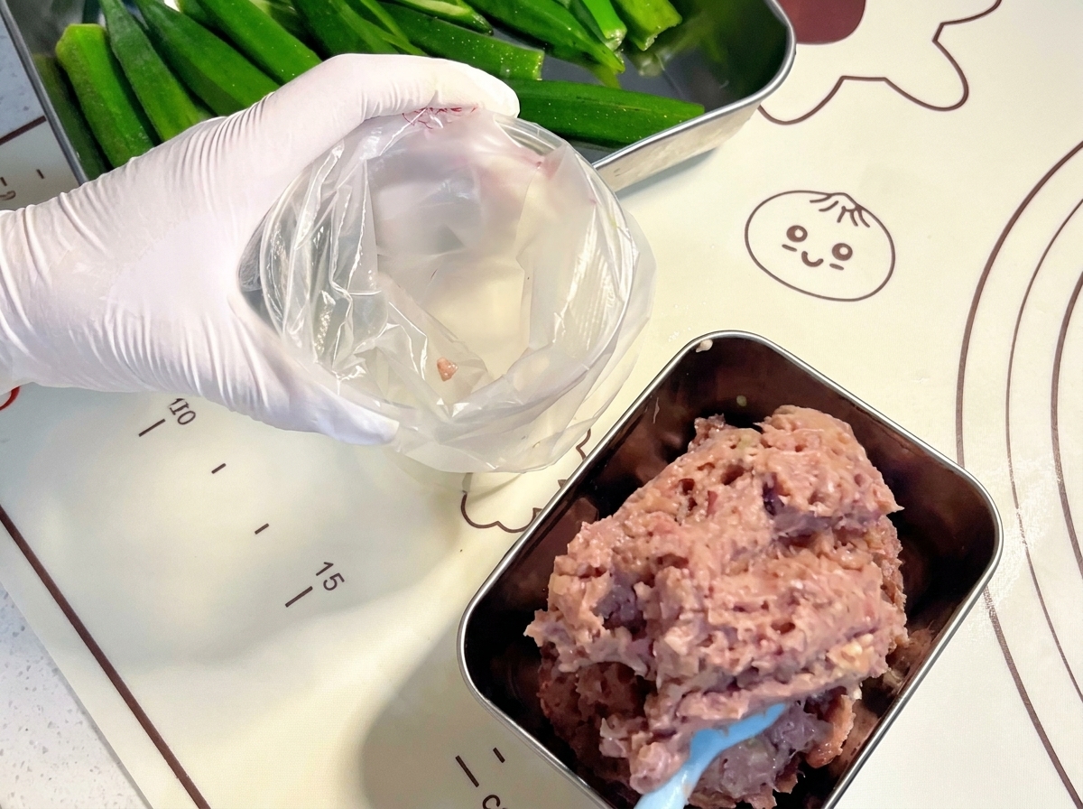 青いスパチュラを使って、混ぜ合わせた生の肉ペーストを透明なプラスチック製の絞り袋に入れている手袋をした手。