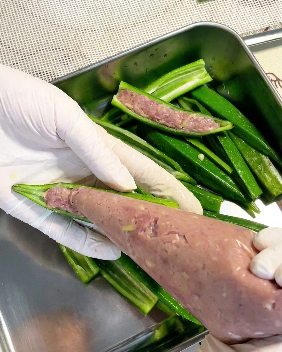透明な絞り袋を使って、下茹でしたオクラの半分に滑らかな淡い色の肉ペーストを詰めている手袋をした手。