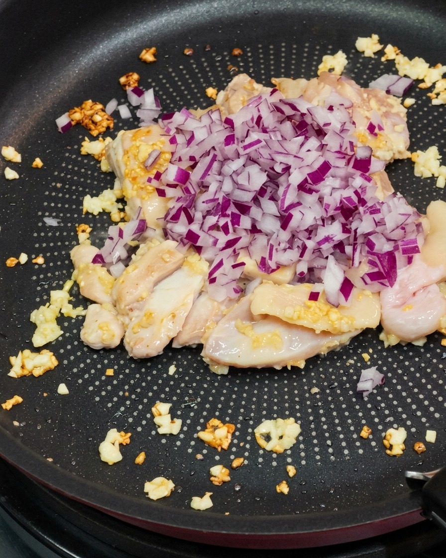 鶏肉とにんにくを炒めたフライパンに紫玉ねぎのみじん切りを加えている様子。