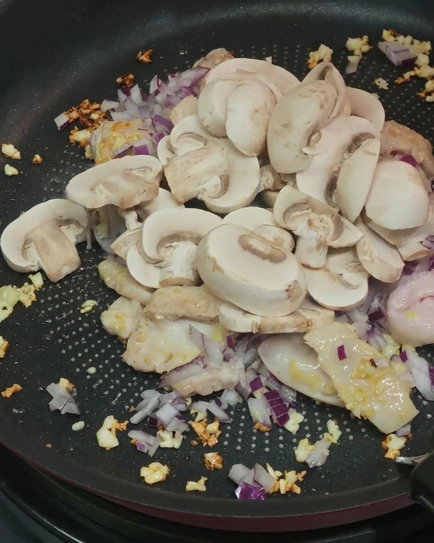 炒めた鶏肉、にんにく、紫玉ねぎが入ったフライパンに、スライスしたホワイトマッシュルームを加えている様子。