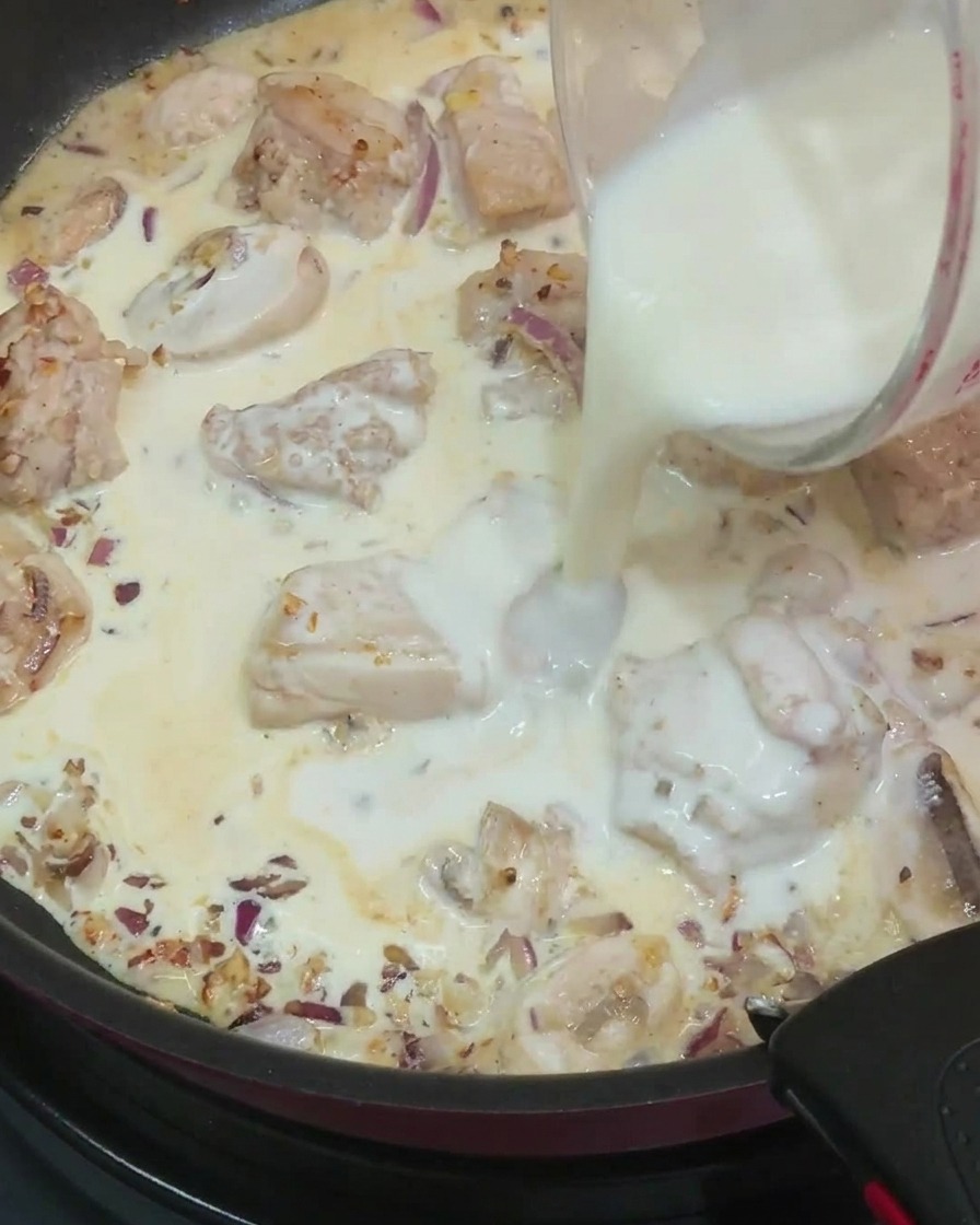 ガラスの計量カップから、炒めた鶏肉、マッシュルーム、玉ねぎ、生クリームが入ったフライパンに牛乳を注いでいる様子。