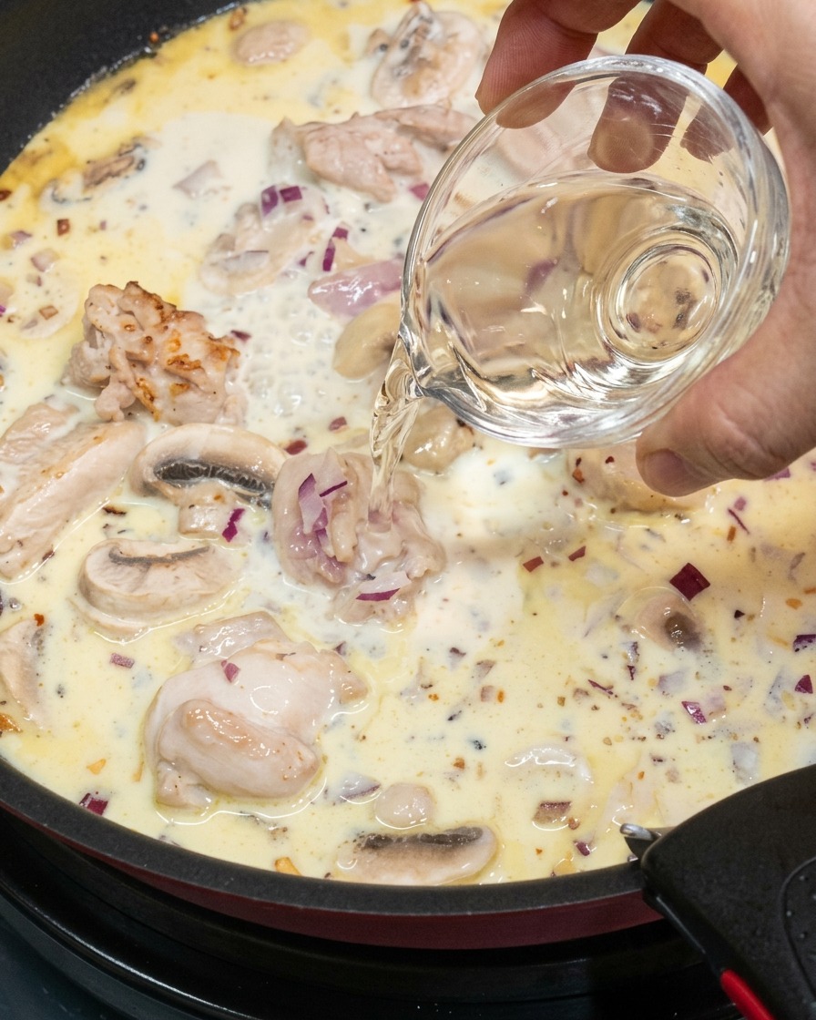 鶏肉とマッシュルームが入ったクリーミーなソースに、小さなガラスのボウルから白ワインを注いでいる様子。