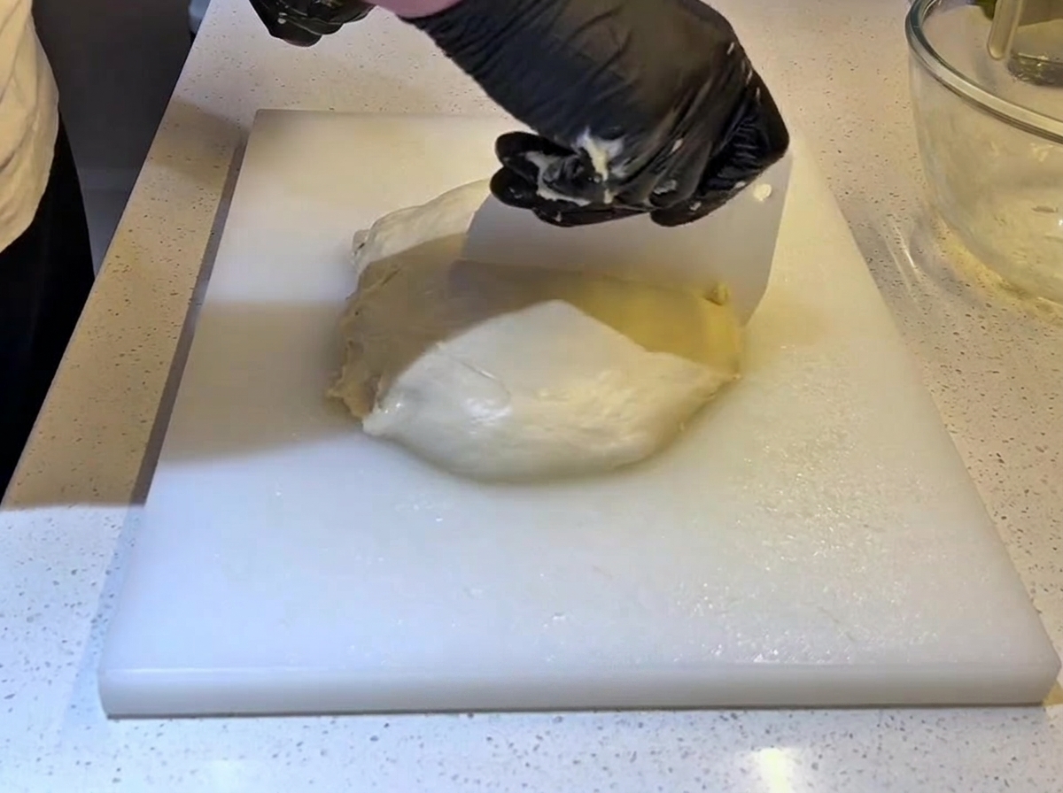 白いプラスチック製のスケッパーを使い、黒い手袋をした手で大きな生地を半分に切り分けている様子。