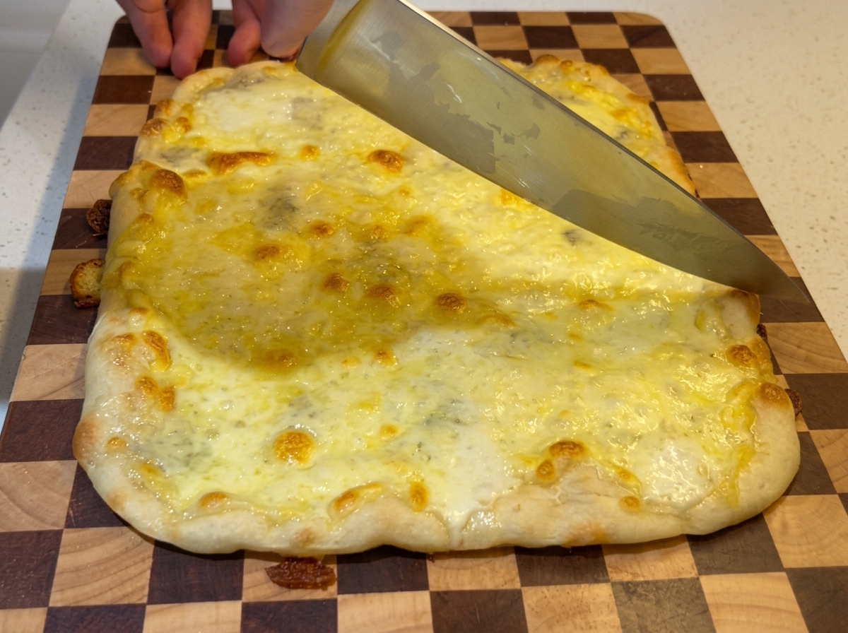 木のまな板に乗せた、チーズがグツグツと煮え立つ焼き立ての長方形ピザを、大きな牛刀で切り分けている様子。