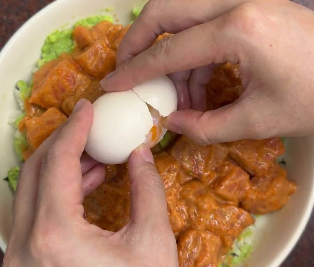 サーモンとアボカドを盛り付けた丼の中央に、両手で温泉卵を優しく割り落としている様子。