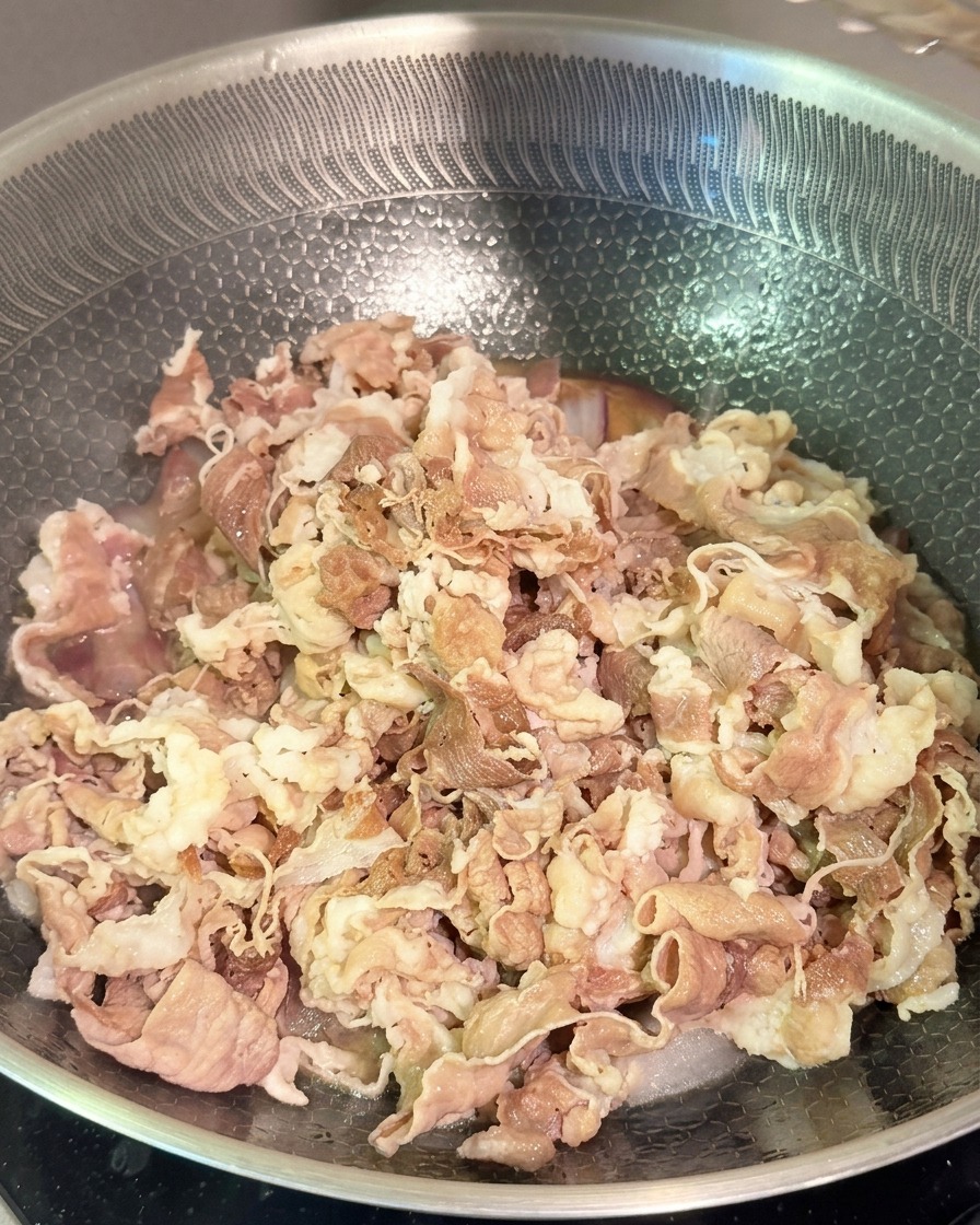 ステンレスのフライパンの中で、柔らかく下茹でした牛肉、赤玉ねぎ、そして濃い色のタレを炒め合わせている様子。