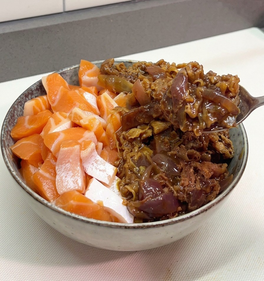 サーモンが盛り付けられた丼のもう半分のスペースに、金属製のスプーンでタレの絡んだ温かい牛肉と赤玉ねぎを盛り付けている様子。