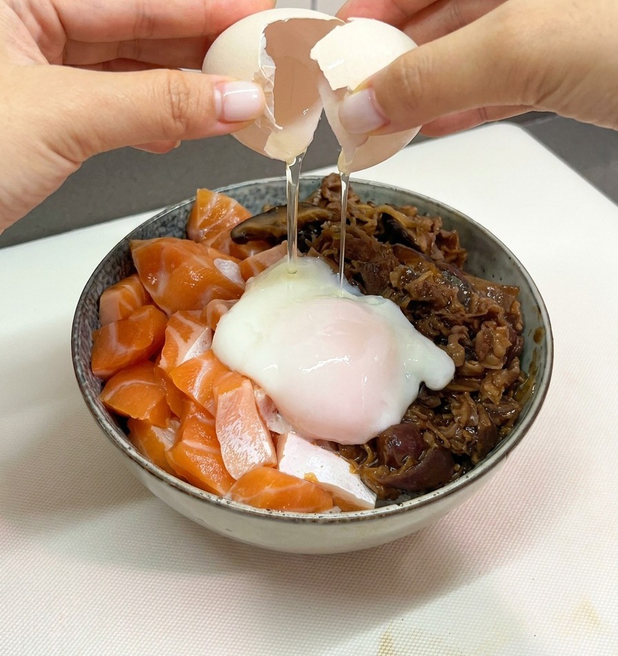 両手で白い卵の殻を割り、サーモンと牛肉がのった丼の中央に、ぷるぷるとした温泉卵を優しく落としている様子。