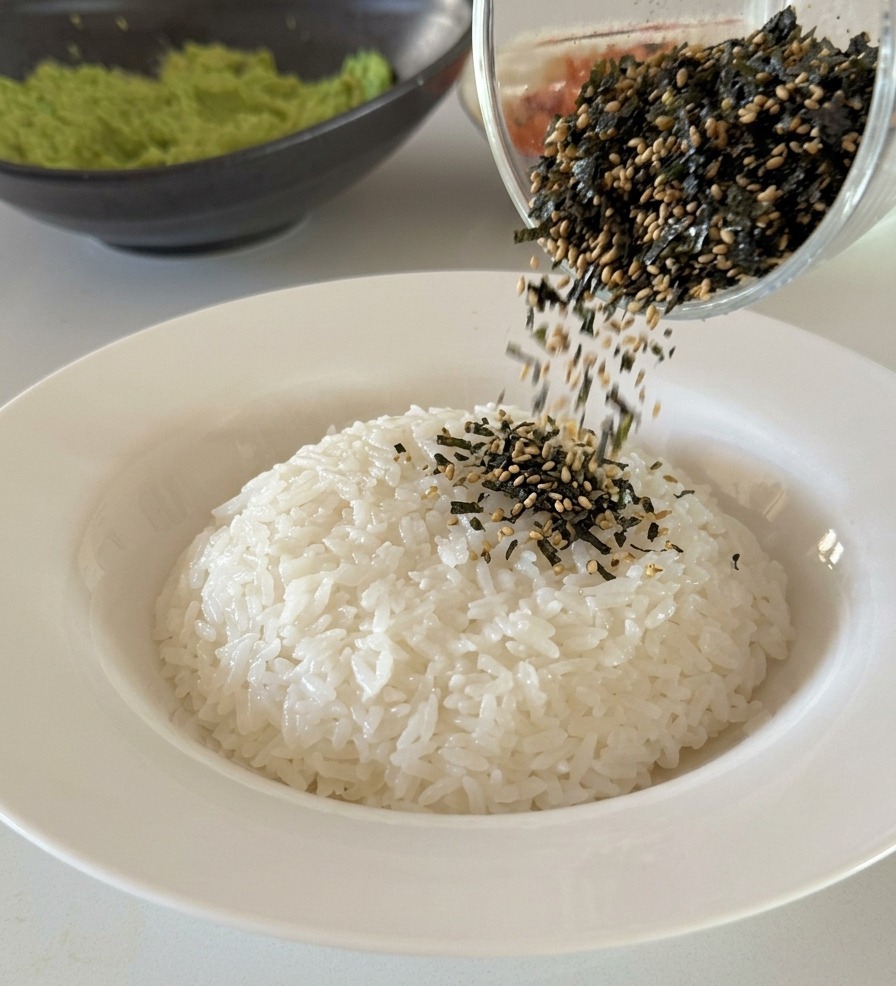 丼に盛られた温かい白ご飯の上に、ガラスの器からたっぷりの刻み海苔と白いりごまを振りかけている様子。
