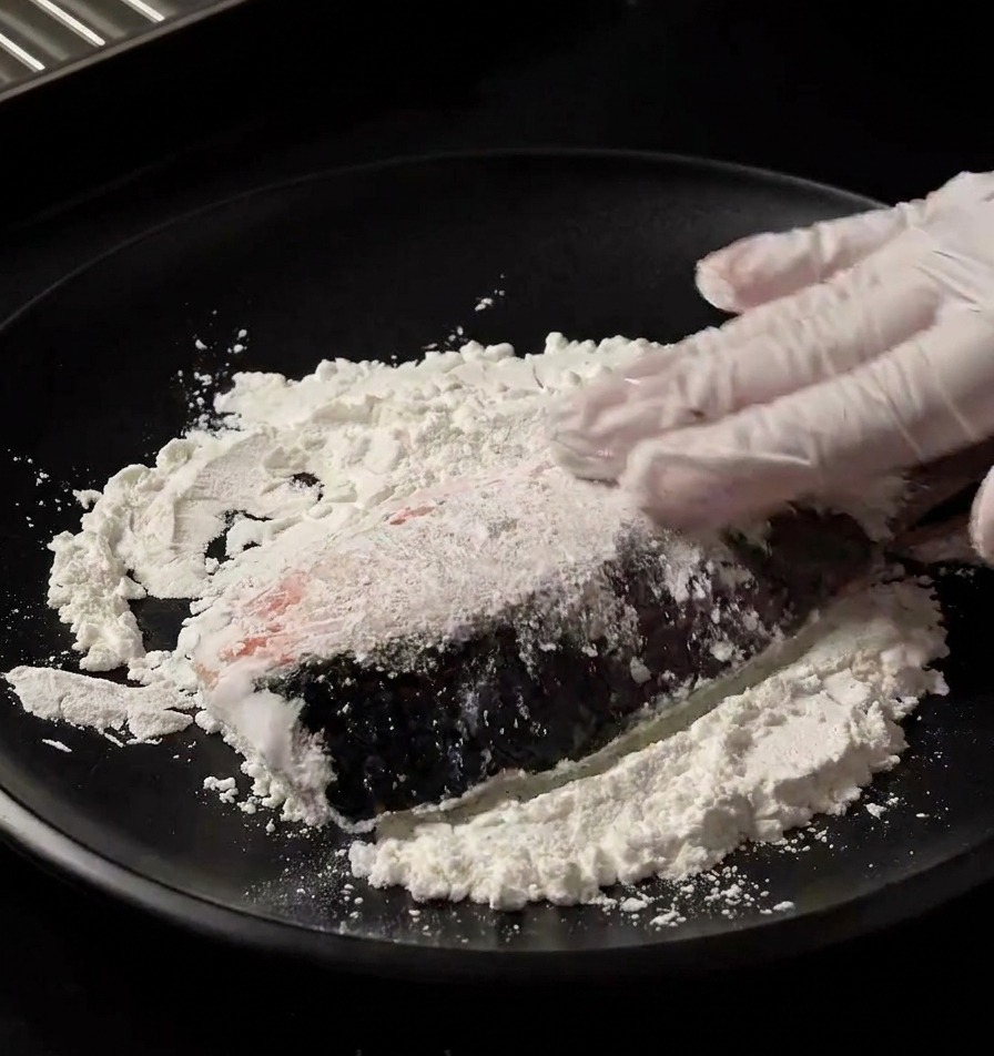 手袋をはめた手で、黒い皿に広げられた白い片栗粉に生の鮭の切り身を押し付け、均一にまぶしている様子。