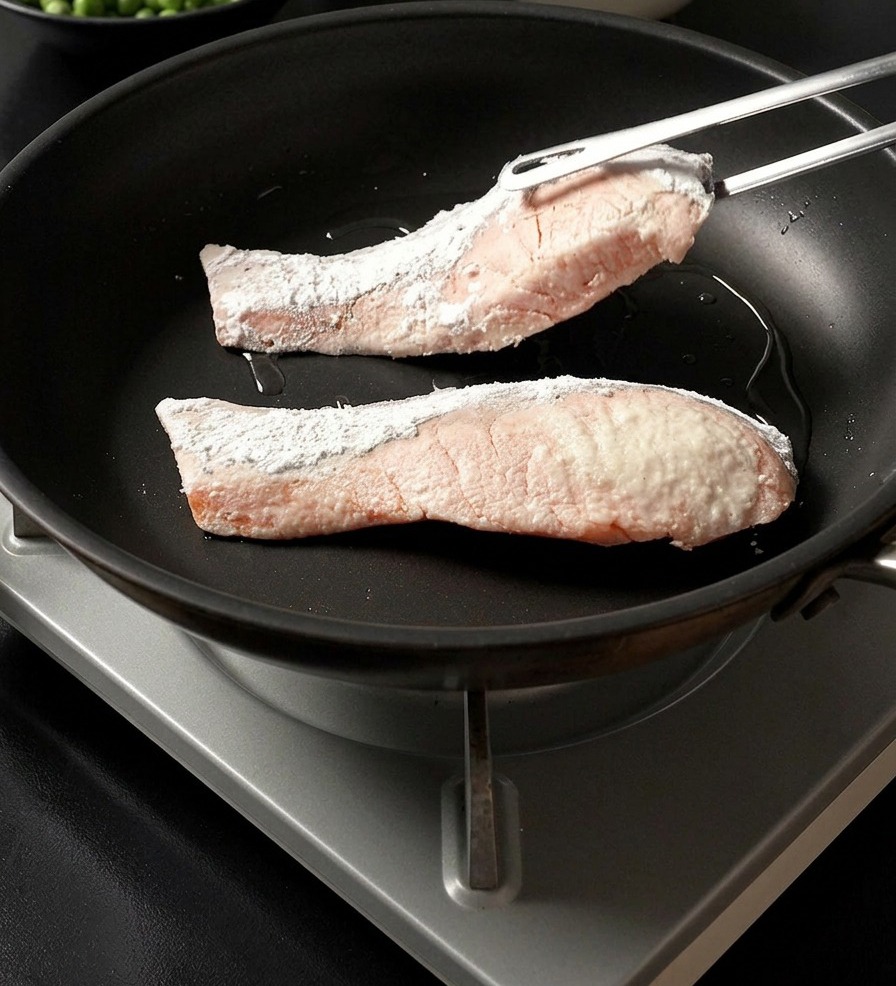金属製のトングを使い、片栗粉をまぶした鮭の切り身を、熱したフライパンに並べて入れている様子。