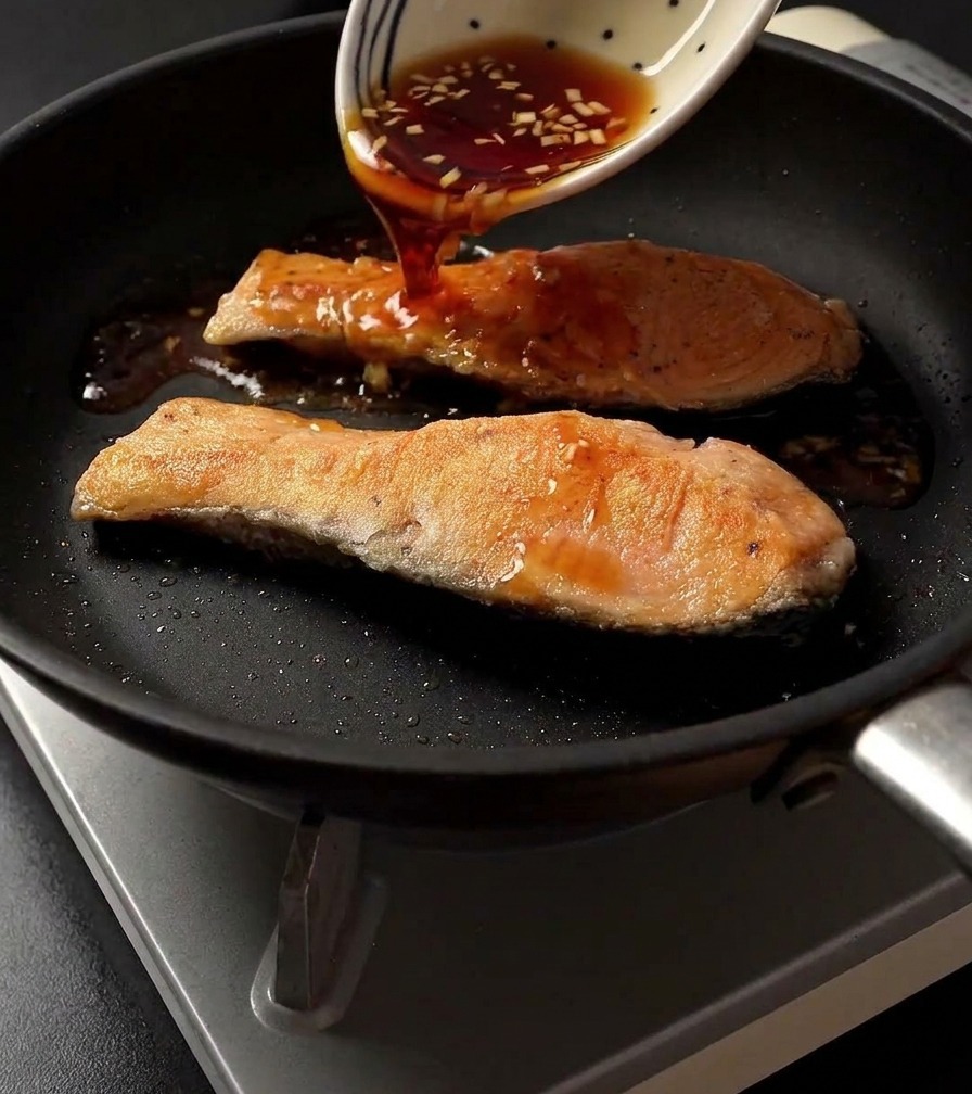 フライパンでこんがりと焼けた2切れの鮭の上から、柄物の白いボウルに入った濃い色の照り焼きタレを注ぎ入れている様子。