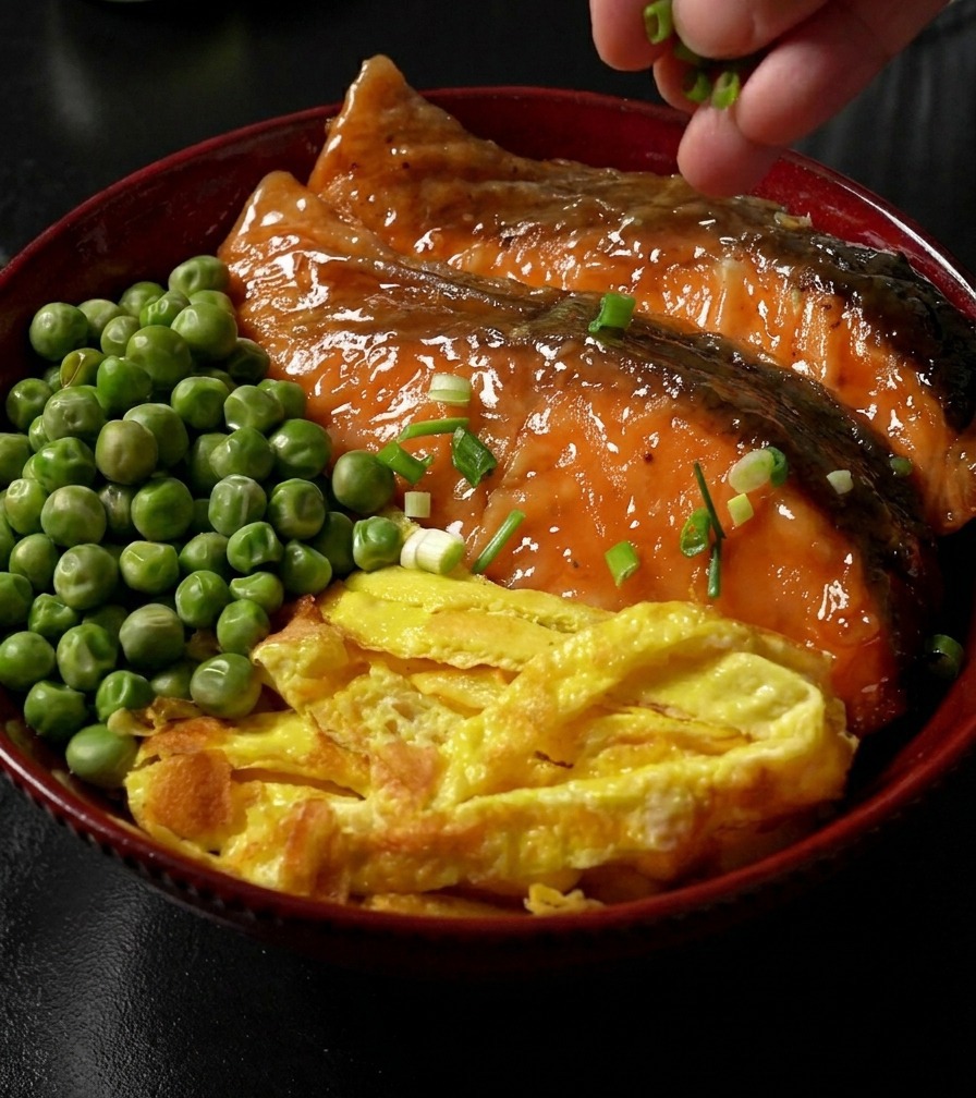 ご飯の上に照り焼き鮭、錦糸卵、グリーンピースを乗せ、仕上げに手で刻み青ねぎを散らしている完成した丼の様子。