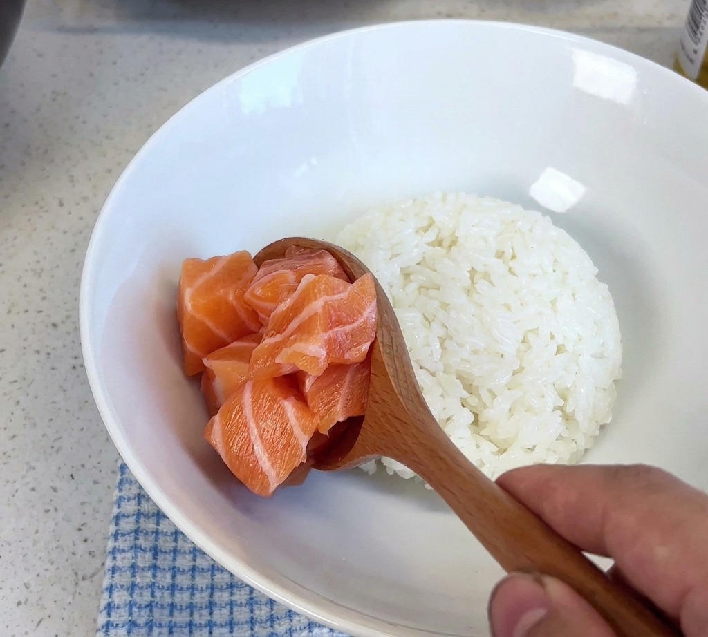 木製のスプーンで角切りサーモンをすくい、白いご飯が入った器に盛り付けている様子