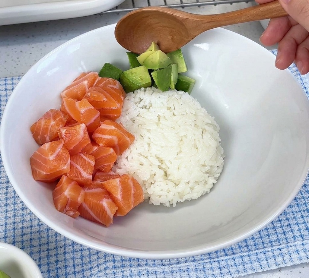 サーモンをのせた白い器のご飯の上に、角切りのアボカドをスプーンで添えている様子