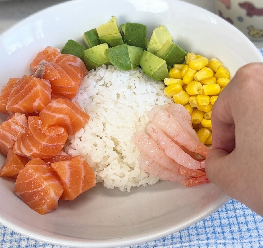サーモンやコーンがのった器のご飯の上に、ピンク色で透明感のある甘エビを盛り付けている手元