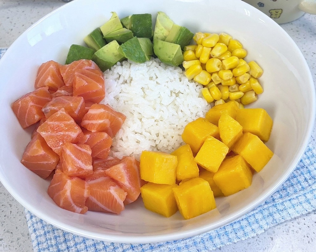 ご飯、サーモン、アボカド、コーン、エビが盛られたカラフルなポキ丼に、鮮やかな黄色のマンゴーが加わった様子