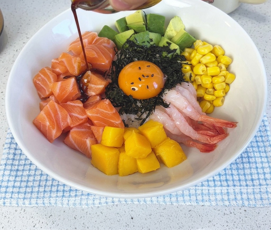 完成したサーモンポキ丼と卵黄の上から、小さなガラス of 器に入った濃いワサビ醤油を回しかけている様子