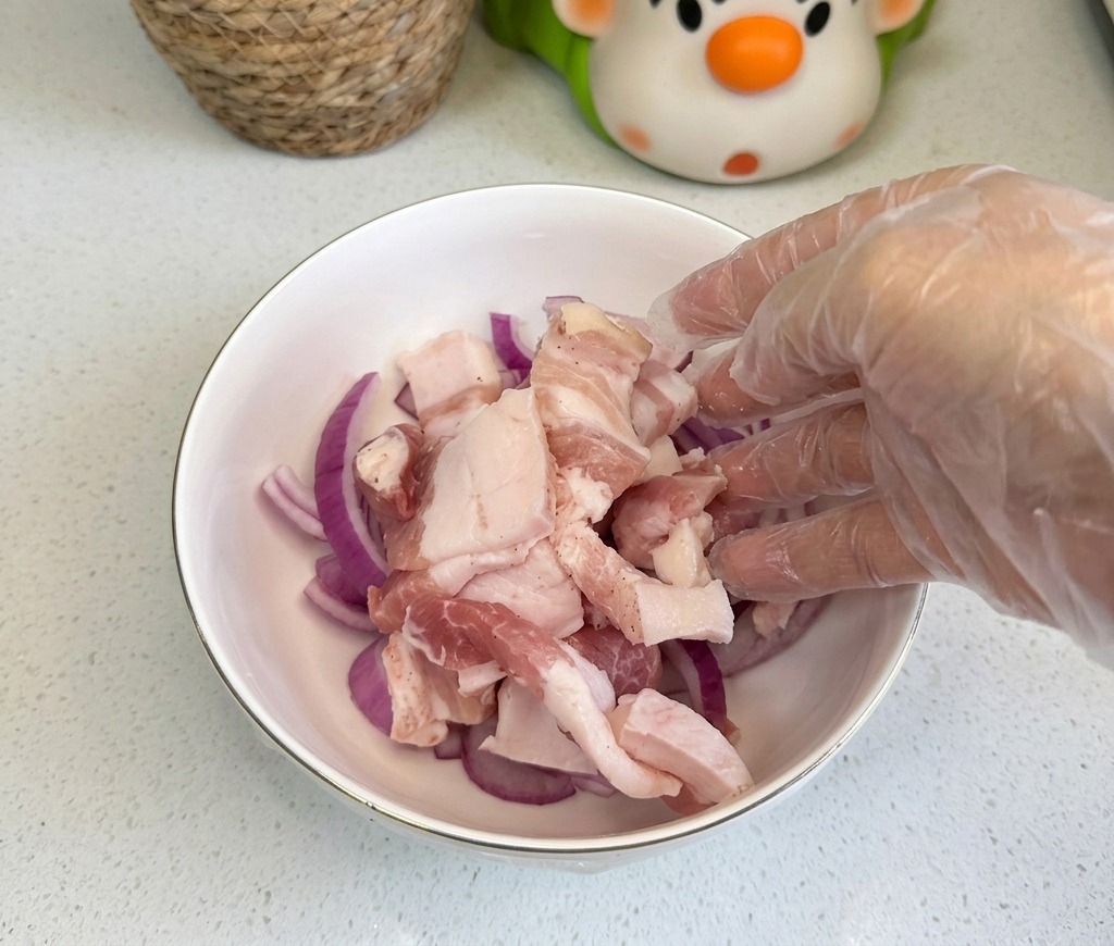 薄切りにした赤玉ねぎが入った白いボウルに、生の豚バラ肉の薄切りを加えている様子。